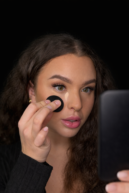 Woman applying makeup with a black SOANERA sponge in front of a mirror on a black background