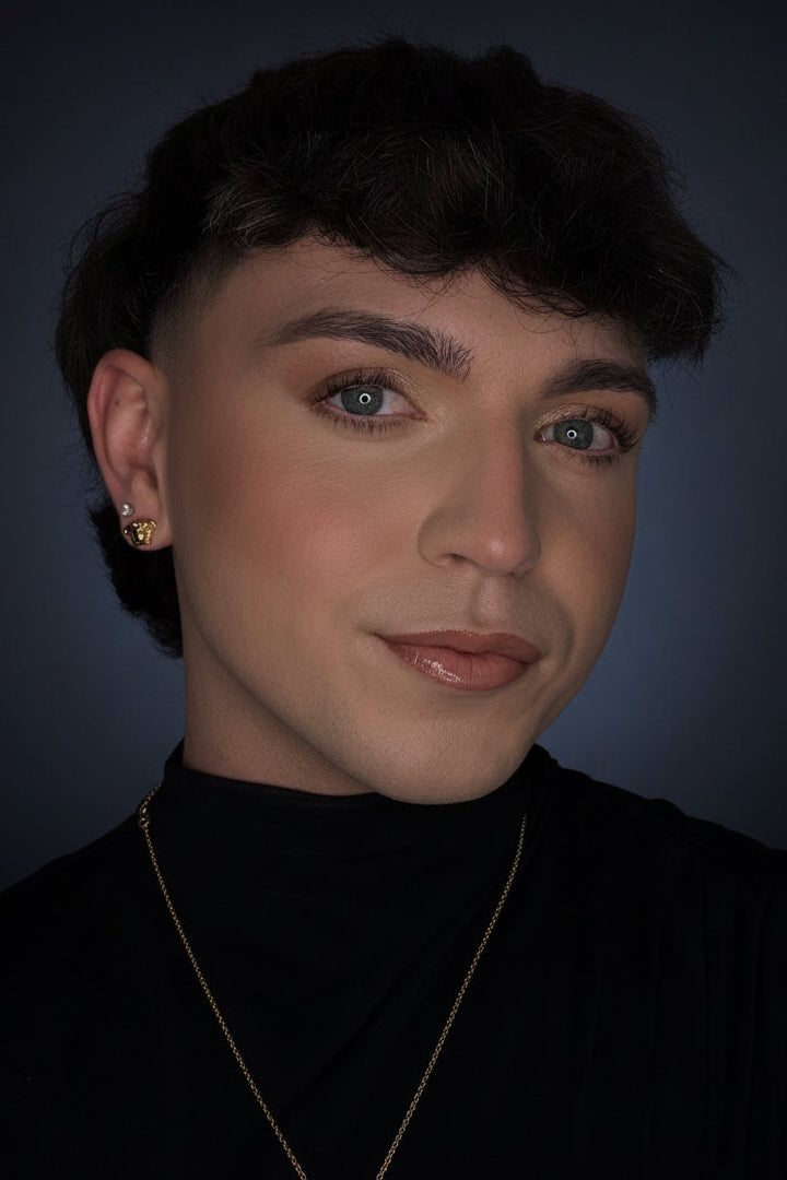A close-up portrait of a person with short dark hair wearing a black turtleneck, a delicate gold chain necklace and a small gold stud earring, set against a dark blue-gray background.